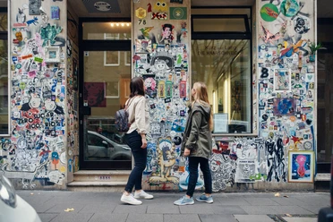 Belgisches Viertel Zwei Personen betrachten eine mit Stickern und Graffiti verzierte Wand in einer Stadtstraße.