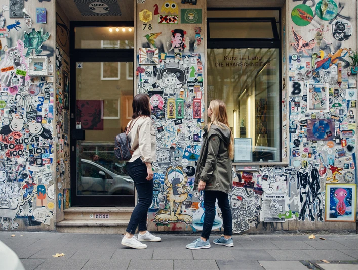 Belgisches Viertel Zwei Personen betrachten eine mit Stickern und Graffiti verzierte Wand in einer Stadtstraße.Two people look at a wall decorated with stickers and graffiti in a city street.
