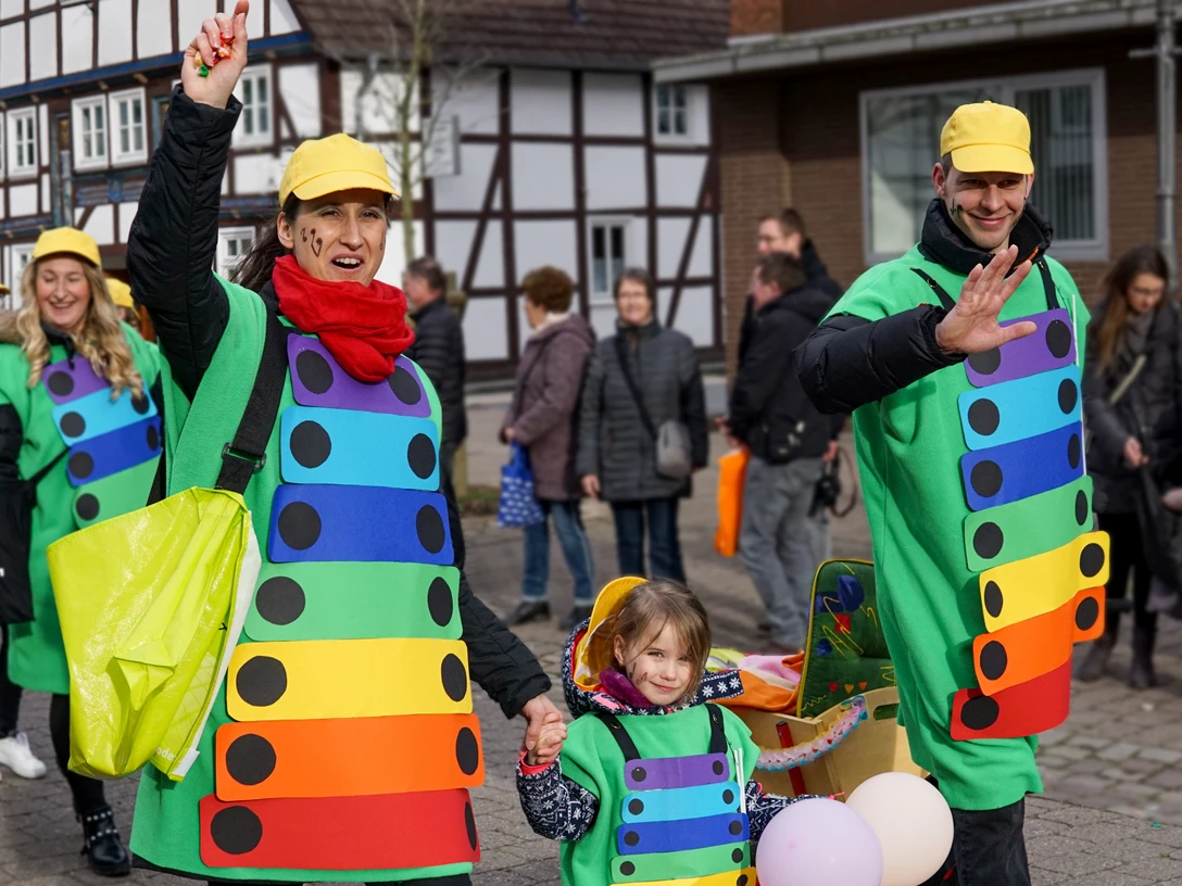 Kindercarnevalsumzug Bild 2 - Sascha Mantel.jpg Menschen in kunterbunten Kostümen mit gelben Hüten bei einem Karnevalsumzug in einer Stadt.