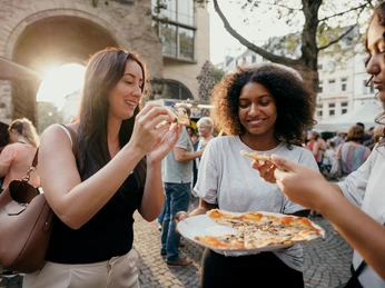 meet & eat Köln Sonnenstrahlen blitzen durch die Bäume, während drei Freundinnen lachend auf einem Markt in Köln Pizza genießen.Rays of sunshine flash through the trees while three friends enjoy pizza at a market in Cologne, laughing.