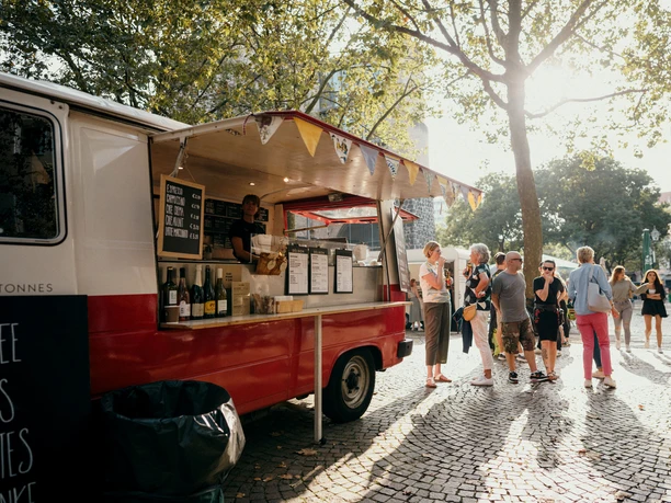 meet & eat Köln Ein farbenfroher Foodtruck auf einem sonnigen Platz, umgeben von Menschen, die in entspannter Atmosphäre Essen genießen.