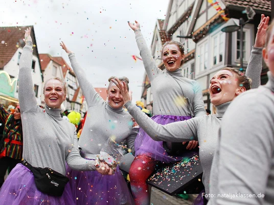 Altweiberumzug Rietberg_2_Foto Natalja Klassen.jpg Gruppe junger Frauen in grauen Oberteilen und lila Röcken wirft buntes Konfetti bei einem Straßenfest.