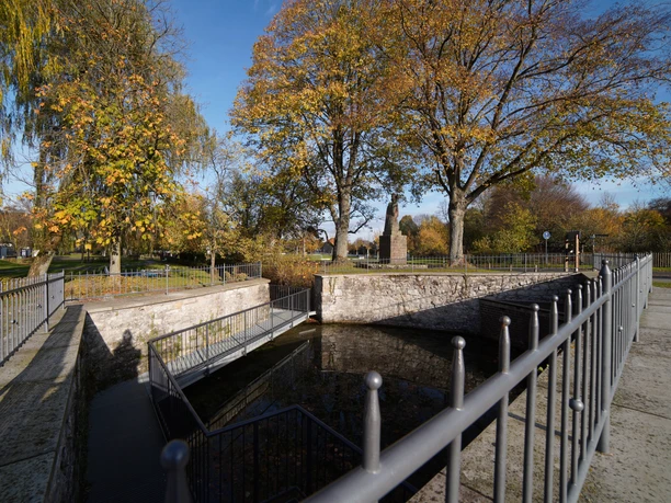 Nikolausquelle Historische Brunnenanlage umgeben von herbstlichen Bäumen und einem Metallgeländer im Freien.