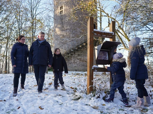 Immer auf der Hut.jpg Eine Familie spaziert im Winterkleid neben einem historischen Steinturm durch den verschneiten Wald.