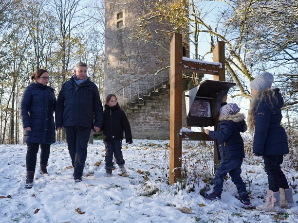 Immer auf der Hut.jpg Eine Familie spaziert im Winterkleid neben einem historischen Steinturm durch den verschneiten Wald.