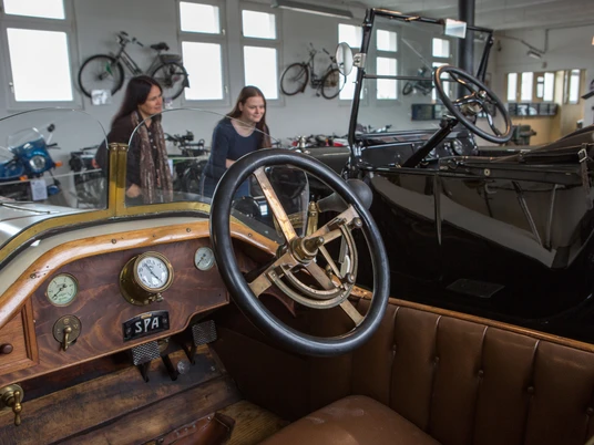 Automuseum Melle Im Automuseum Melle sind historische Autos in einem hellen Raum ausgestellt. Menschen betrachten die Fahrzeuge.