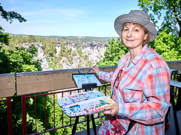 Een vrouw met een hoed schildert op een ezel in een groen, rotsachtig landschap. Ze draagt een kleurrijk geruit overhemd en houdt een kleurenpalet vast.
