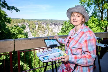 Malkurs an der Bastei mit Andrea Moliére Eine Frau mit Hut malt auf einer Staffelei in einer grünen, felsigen Landschaft. Sie trägt ein bunt kariertes Hemd und hält eine Farbpalette.