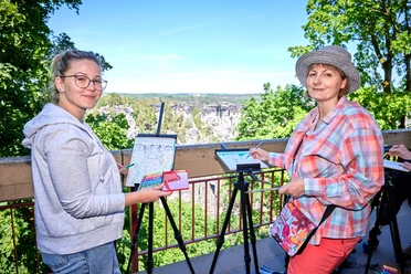 Two women are painting on easels in a green, sunny landscape. One is wearing a gray jacket, the other a colorful plaid shirt and a hat.