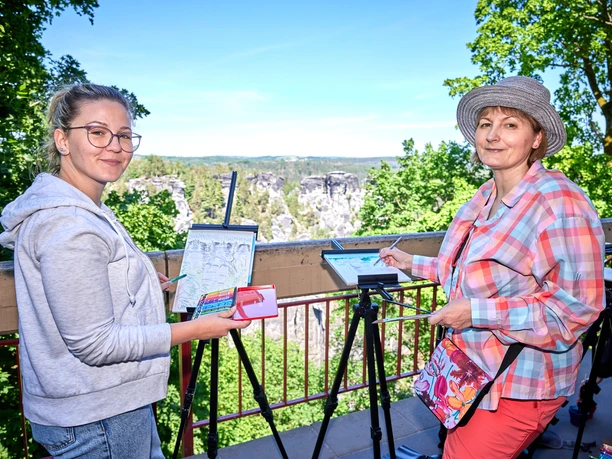 Malkurs an der Bastei mit Andrea Moliére Zwei Frauen malen auf Staffeleien in einer grünen, sonnigen Landschaft. Eine trägt eine graue Jacke, die andere ein bunt kariertes Hemd und einen Hut.
