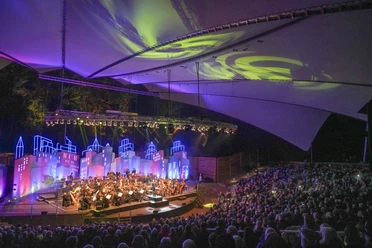 filmmeetsclassic22_57.jpg Orchester spielt vor farbig beleuchteter Kulisse auf der Freilichtbühne Meppen unter Zeltdach.