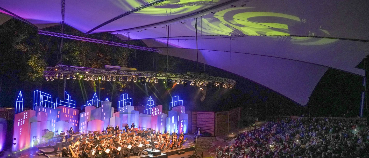 filmmeetsclassic22_57.jpg Orchester spielt vor farbig beleuchteter Kulisse auf der Freilichtbühne Meppen unter Zeltdach.