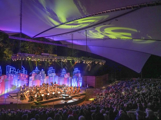 filmmeetsclassic22_57.jpg Orchester spielt vor farbig beleuchteter Kulisse auf der Freilichtbühne Meppen unter Zeltdach.