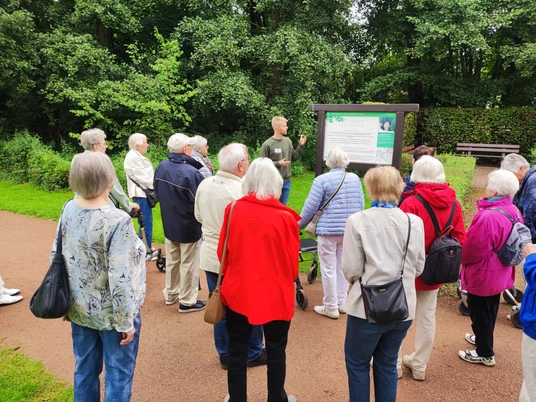 Kurparkführung Eine Gruppe von Senioren lauscht einem Guide bei einer Führung durch einen grünen Park.