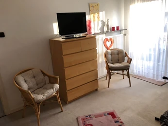 Wohnzimmer Kuhls Helles Wohnzimmer mit Kommode, kleinem Flachbildfernseher und zwei Korbstühlen vor dem Fenster.