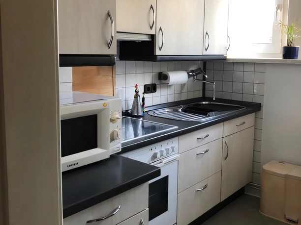 Küche Kuhls Helle, funktionale Einbauküche mit Herd, Mikrowelle, Spüle und weißen Fronten unter Tageslicht.