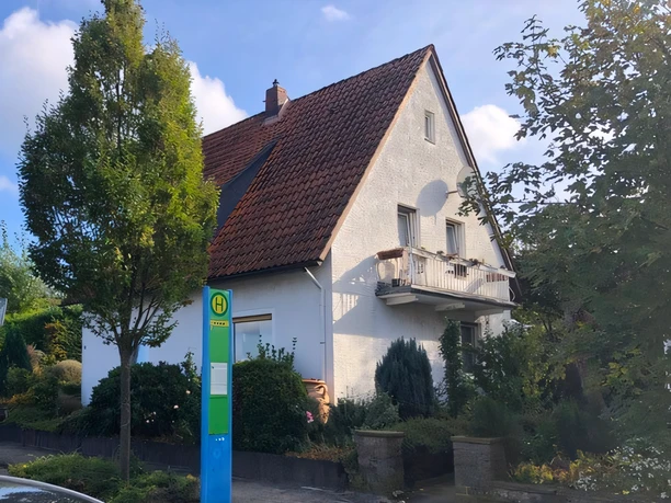 Außenansicht Kuhls Zweigeschossiges weißes Haus mit rotem Ziegeldach, Balkon, umgeben von Bäumen, nahe Bushaltestelle.