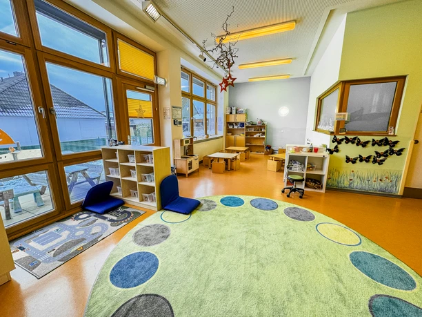 Kinderkrippe-Amelinghausen-03.jpg Heller Gruppenraum der Kinderkrippe Amelinghausen mit rundem Teppich und Spielbereichen.