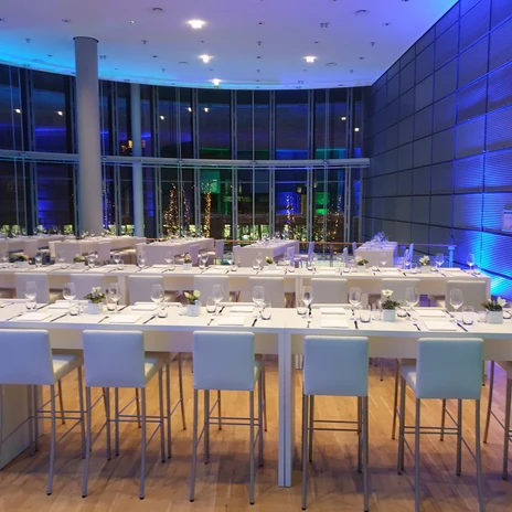 Asta Galerie Heller Raum mit hohen Fenstern, festlich gedeckten weiĂźen Tischen und blauer Lichtbeleuchtung.