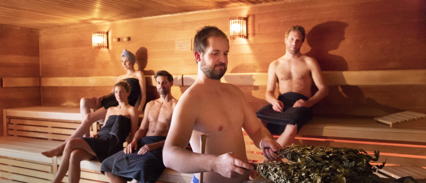 Koppke - Das neue Saunabad in Greetsiel Mehrere Personen entspannen in einer modernen Holzsauna, während ein Saunameister einen Aufguss vorbereitet.