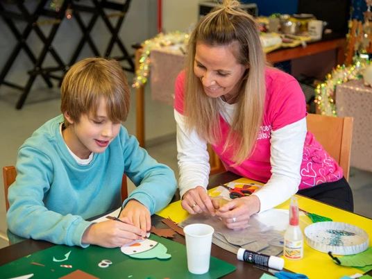 Lükko-Leuchtturm-Kinderhaus Greetsiel Auf dem Bild sieht man einen kleinen Jungen zusammen mit Anke vom Kinderhaus am Tisch sitzend basteln.