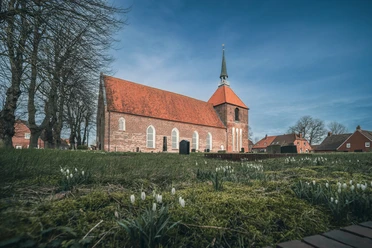 Rysum Kirche Backsteinkirche mit rotem Ziegeldach und Turm im Rundwarfendorf Rysum, umgeben von alten Bäumen.