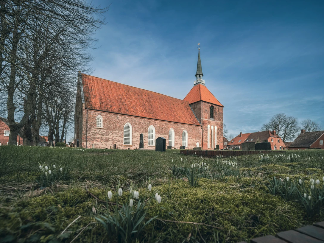 Rysum Kirche Backsteinkirche mit rotem Ziegeldach und Turm im Rundwarfendorf Rysum, umgeben von alten Bäumen.