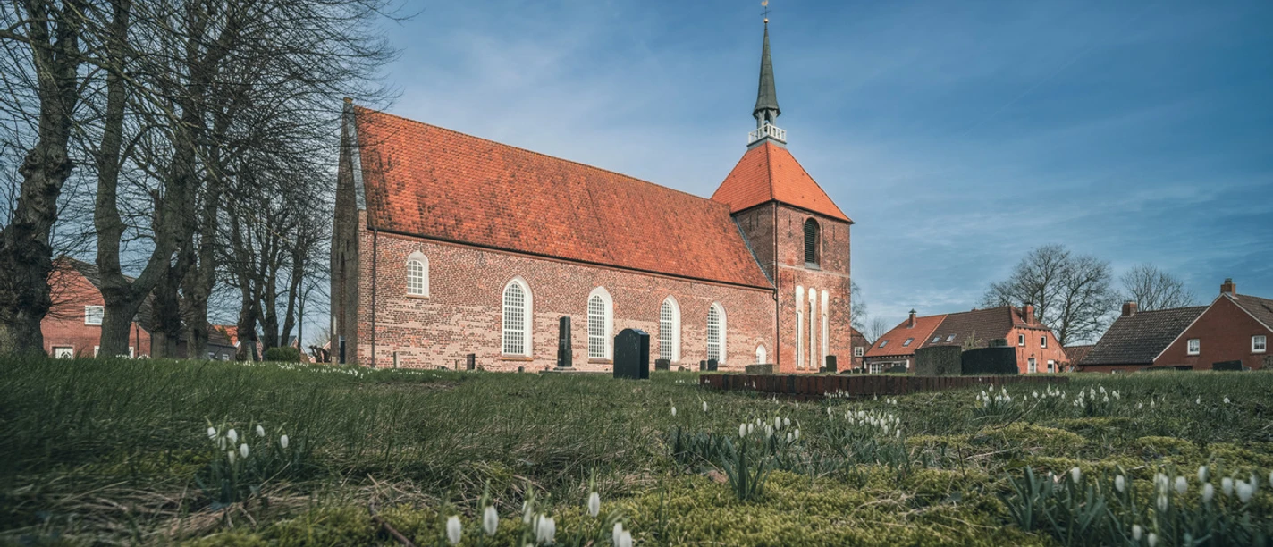 Rysum Kirche Backsteinkirche mit rotem Ziegeldach und Turm im Rundwarfendorf Rysum, umgeben von alten Bäumen.
