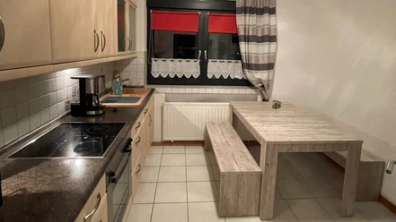 Offene, helle Küche mit modernen Elektrogeräten, Tisch mit Holzbank, Blick auf ein Fenster mit Vorhängen.