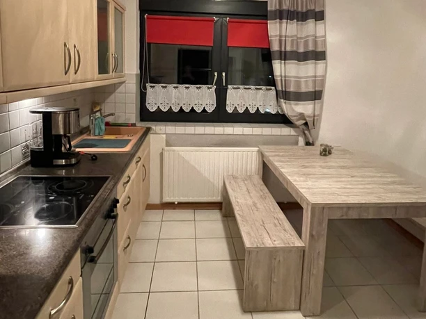 Offene, helle Küche mit modernen Elektrogeräten, Tisch mit Holzbank, Blick auf ein Fenster mit Vorhängen.