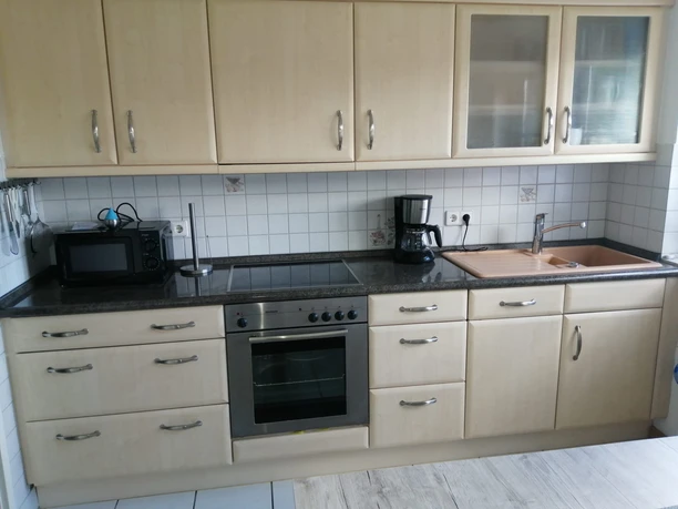 Helle Holzküchenfront mit Glashängeschränken, Elektroherd, Spüle, Kaffeemaschine und Mikrowelle.