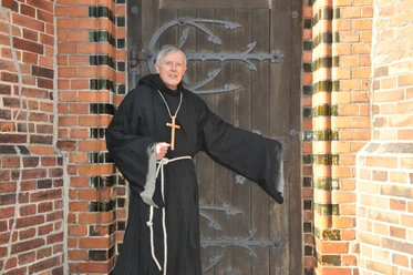 Klosterbruder Jakob steht mit Holzkreuz vor einer historischen Holztür.
