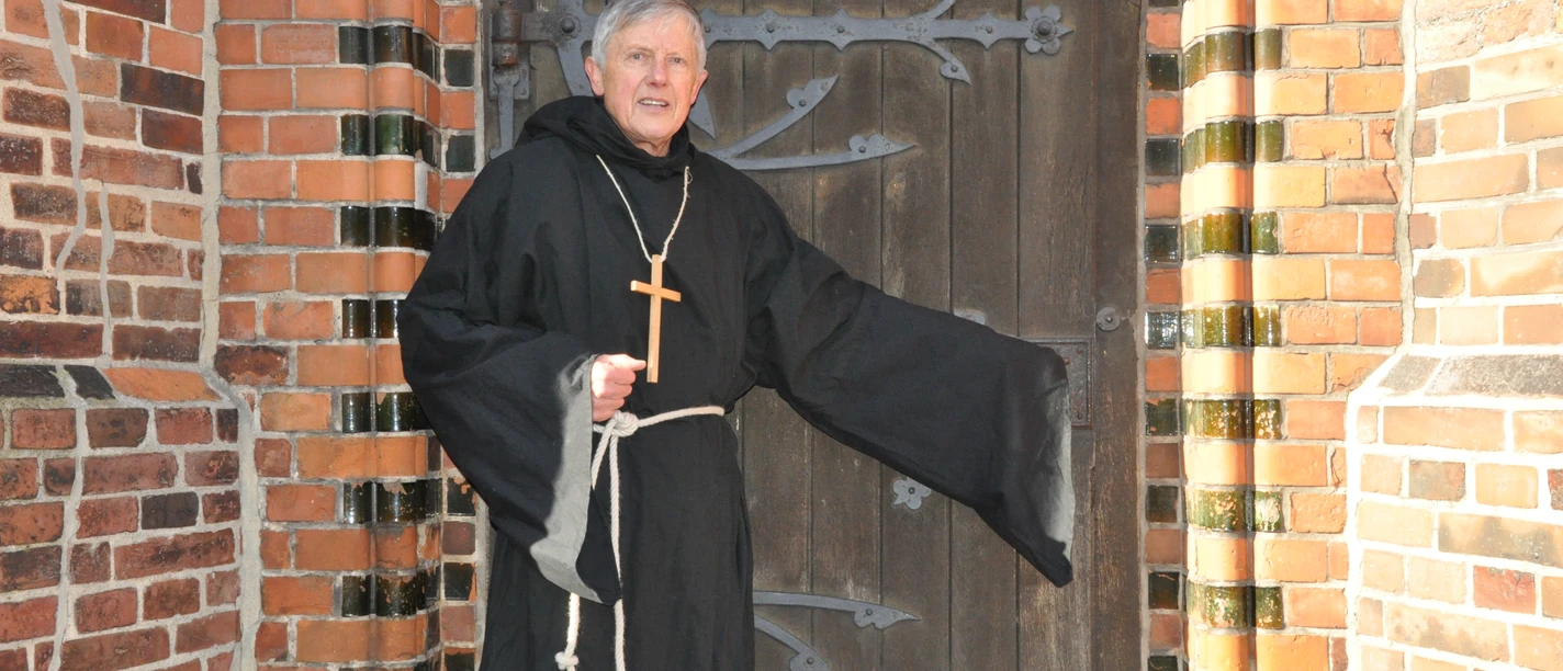 Klosterbruder Jakob steht mit Holzkreuz vor einer historischen Holztür.
