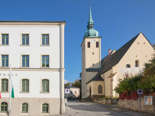 Kostel svatého Petra a Pavla Sebnitz Bílá budova radnice se zelenými okenními rámy a vedle ní kostel se zelenou věží na pozadí modré oblohy.