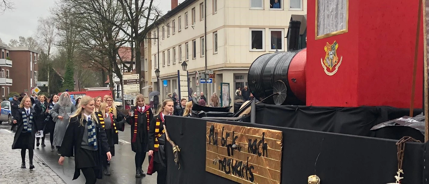 Harry Potter 3.jpg Menschen in Zauberkostümen laufen neben einem festlich dekorierten Umzugswagen mit Hogwarts-Motiv.