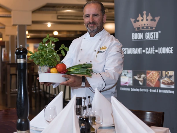 Mehmet_Oerser_c_AngelavonBrill_12.jpg Ein Koch in weißer Uniform präsentiert stolz einen Teller mit frischem Gemüse in einem Restaurant.
