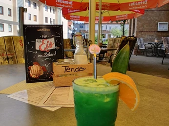 Cocktail Café Tender Grüner Cocktail im Vordergrund mit Zitrusscheiben, Terrasse mit roten Schirmen und Sitzmöglichkeiten hinten.