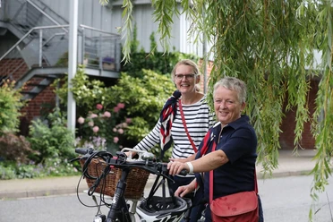 Radtour_Rote_Rosen.jpg Zwei lächelnde Frauen mit Fahrrädern vor einem Backsteingebäude, umrahmt von grünen Weidenzweigen. Geführte Radtour Rote Rosen