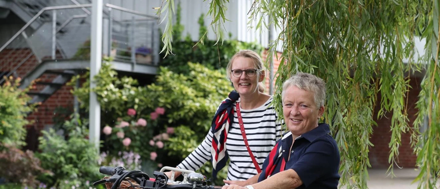 Radtour_Rote_Rosen.jpg Zwei lächelnde Frauen mit Fahrrädern vor einem Backsteingebäude, umrahmt von grünen Weidenzweigen. Geführte Radtour Rote Rosen