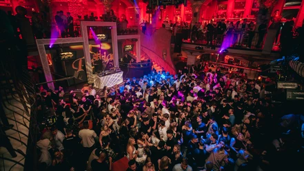 Alando Palais(c)next-choice 4.jpg Menschen tanzen in einem belebten, farbenfroh beleuchteten Nachtclub mit DJ auf einer erhöhten Bühne.