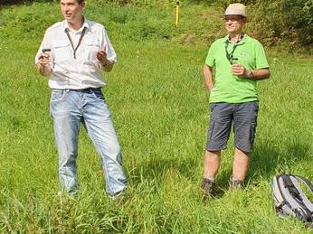 Wolfgang Kaiser (Wega Whisky) und Markus Rösner Zwei Männer stehen in einem grünen Feld vor bewaldetem Hintergrund und scheinen zu diskutieren.
