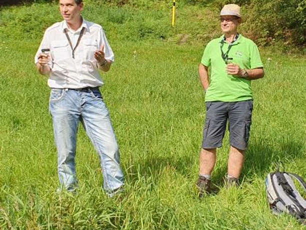 Wolfgang Kaiser (Wega Whisky) und Markus Rösner Zwei Männer stehen in einem grünen Feld vor bewaldetem Hintergrund und scheinen zu diskutieren.