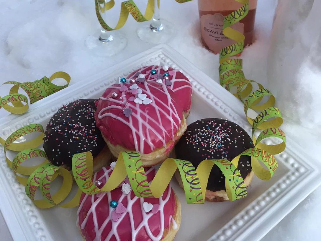 Karneval im Schnee Eine Platte mit Donuts und Sektflasche, dekoriert mit bunten Luftschlangen im Schnee.