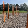 Calisthenics_Sportgeräte am Hahnentanger See Outdoor-Fitnessbereich mit orangen Stangen und Klettergerüsten auf Kiesboden, umgeben von Wiesen und Bäumen