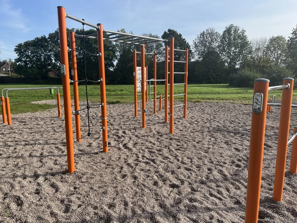 Calisthenics_Sportgeräte am Hahnentanger See Outdoor-Fitnessbereich mit orangen Stangen und Klettergerüsten auf Kiesboden, umgeben von Wiesen und Bäumen