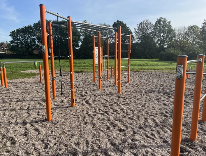Calisthenics_Sportgeräte_IMG_0778.JPG Outdoor-Fitnessbereich mit orangen Stangen und Klettergerüsten auf Kiesboden, umgeben von Wiesen und Bäumen