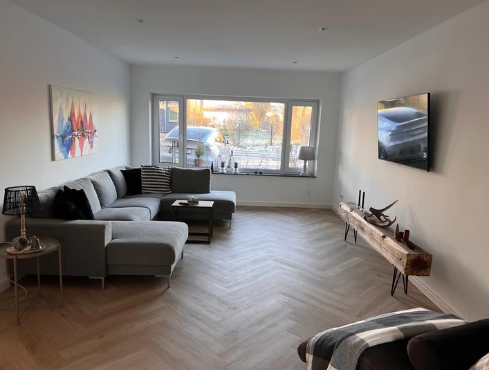 Moderner Wohnbereich mit Ecksofa, TV und großem Fenster, das viel Tageslicht hineinlässt.