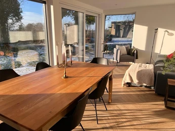 Wintergarten Ein sonnendurchfluteter Wintergarten mit Holztisch, modernen Stühlen und Blick in die verschneite Natur.