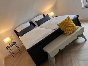 Doppelbett mit blauer und weißer Bettwäsche, gelbes Kissen auf weißer Bank, zwei Nachttischlampen.