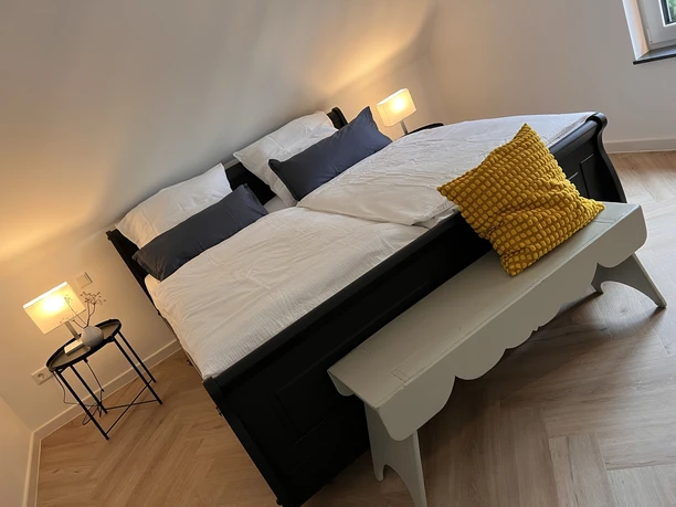 Schlafbereich Doppelbett mit blauer und weißer Bettwäsche, gelbes Kissen auf weißer Bank, zwei Nachttischlampen.